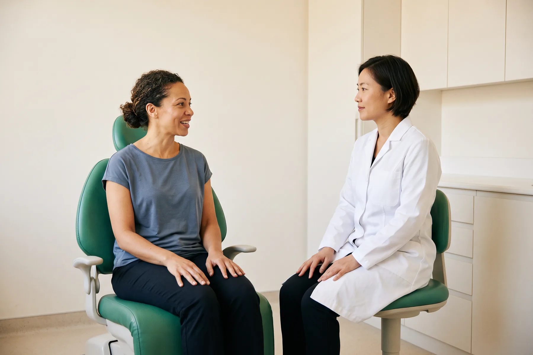 A woman with curly hair tied back and a slate-blue top sits facing a dentist with a dark bob and plain white coat, with both women resting their hands on their laps.