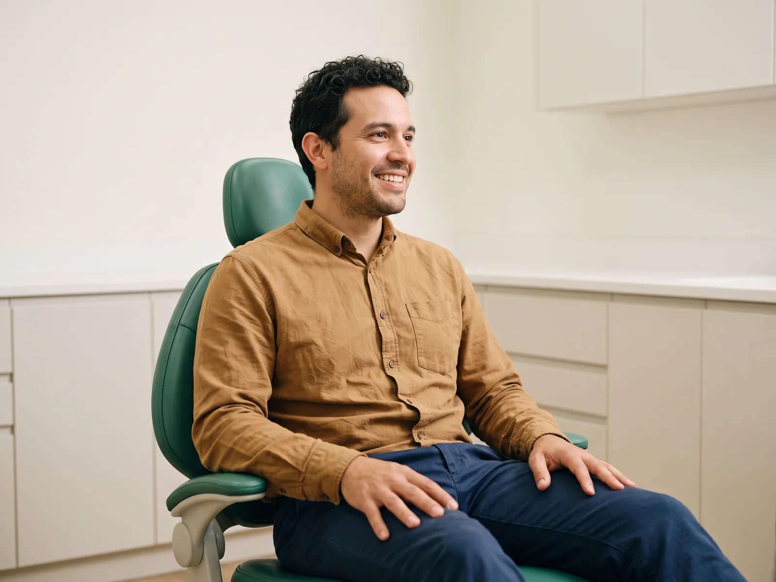 Smiling man with dark curls and a tan shirt seated in a green dental chair