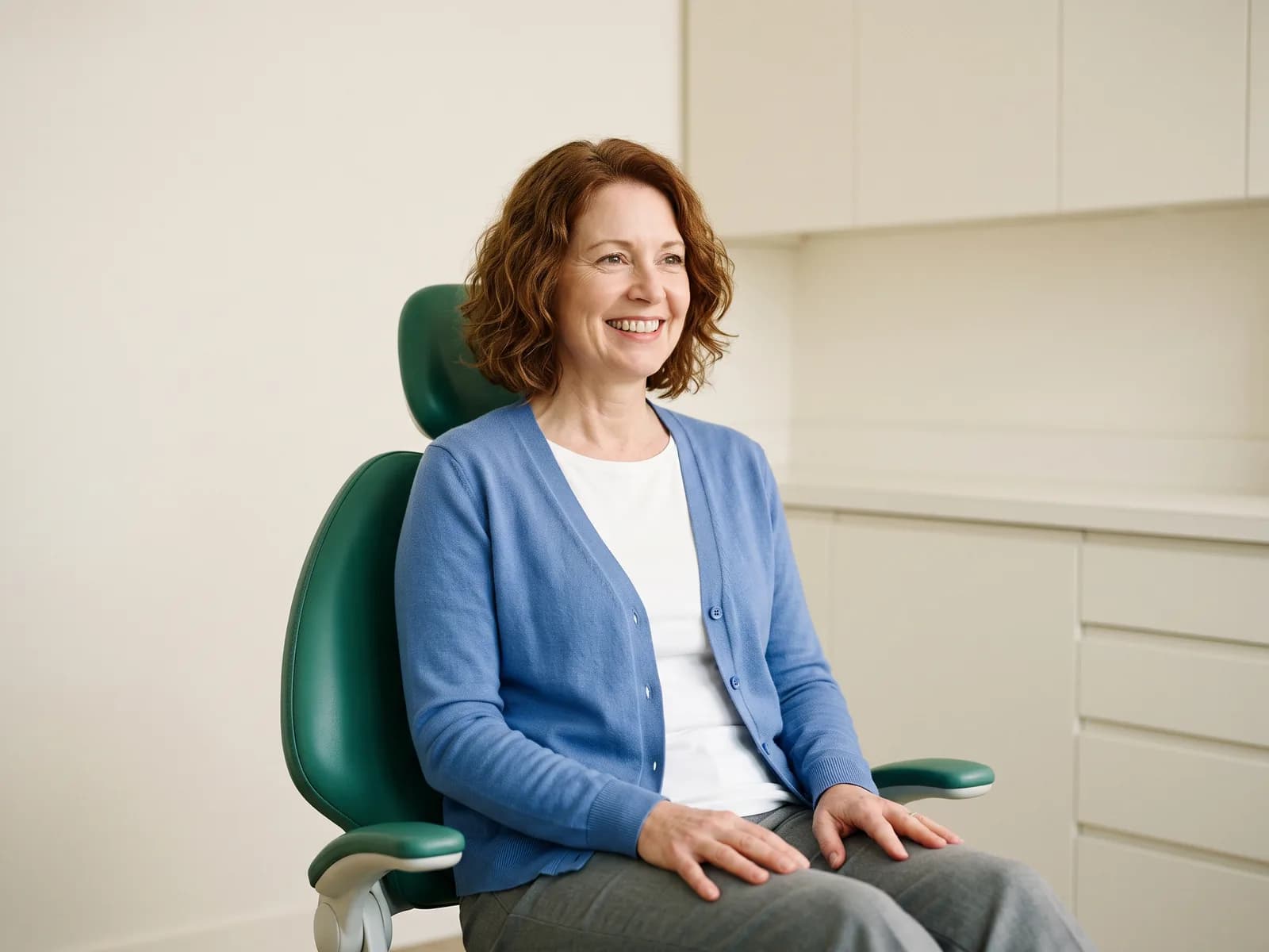 Smiling woman with wavy auburn hair and a blue cardigan seated in a green dental chair