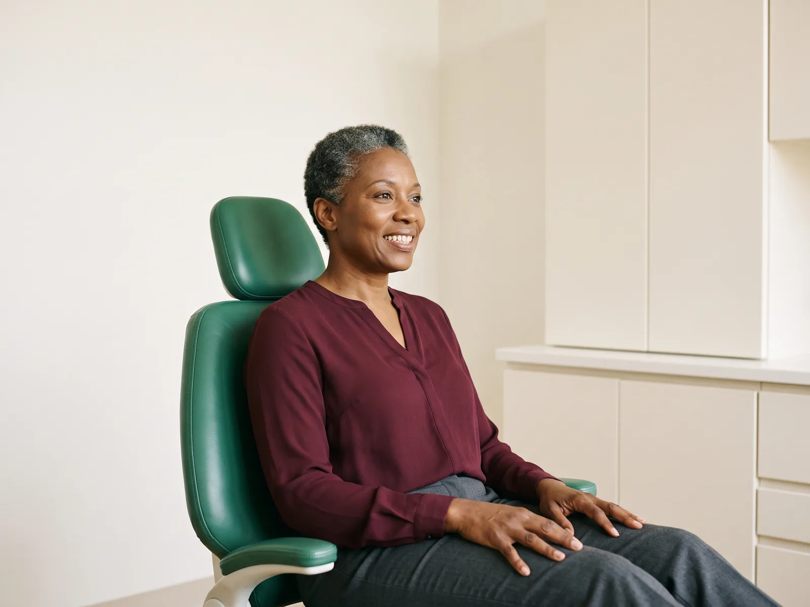 Smiling woman with short gray curls and a burgundy blouse seated in a green dental chair