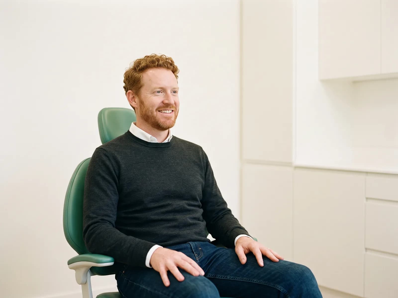 Man with curly red hair and a red beard seated in a green dental chair, wearing a charcoal sweater.