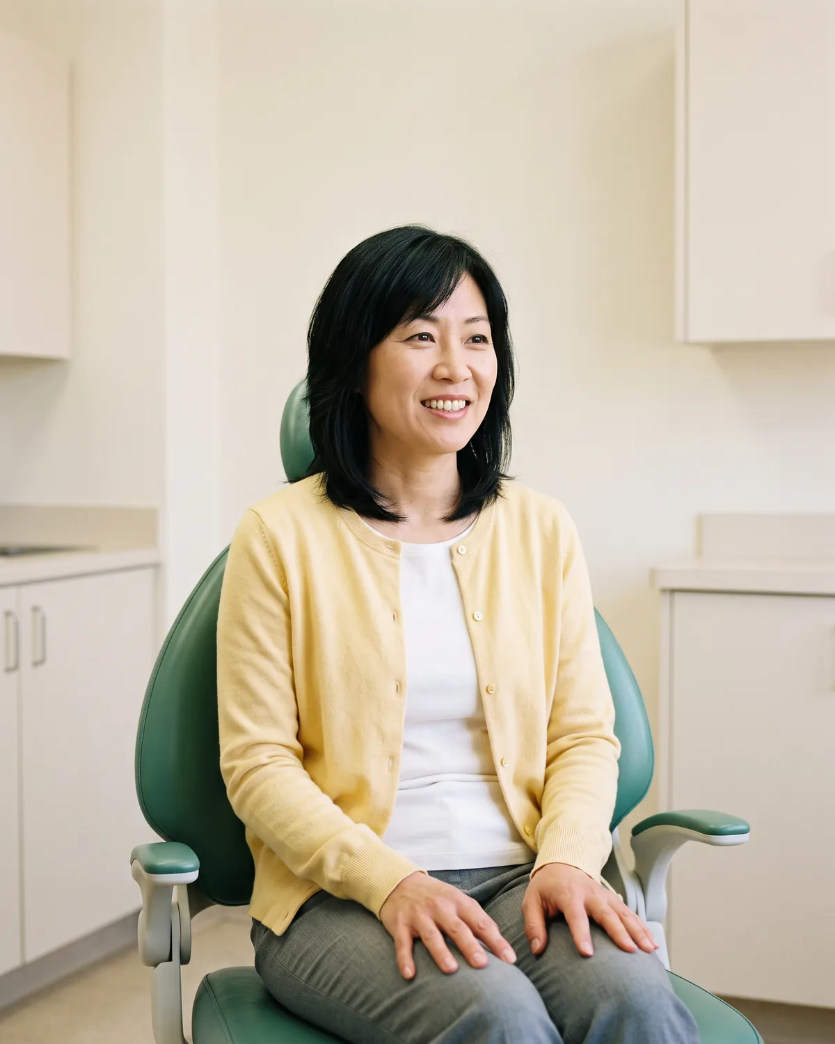 Woman in a yellow cardigan seated in a green dental chair with her hands resting on gray trousers.