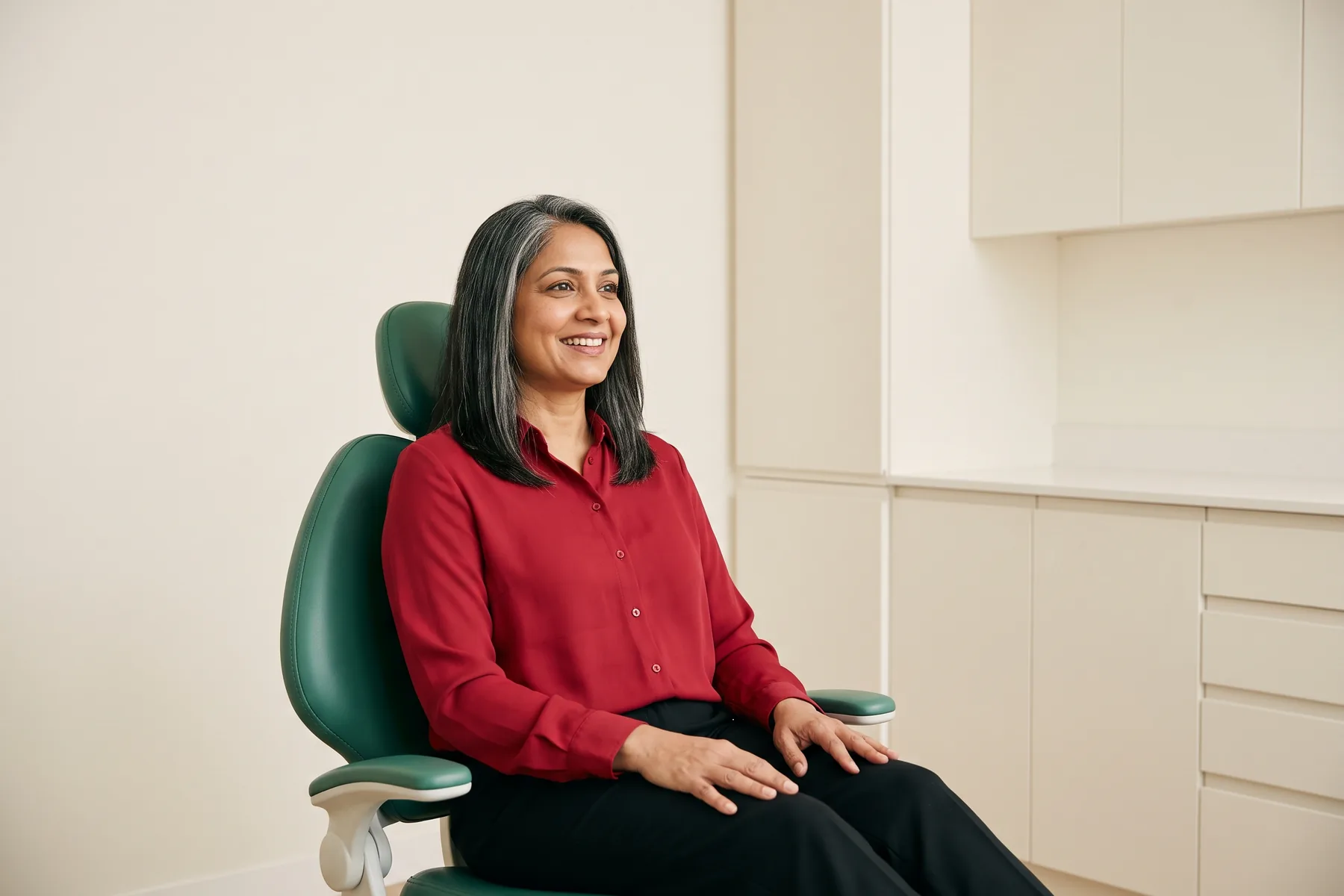 Woman with gray streaks in her dark hair and a red blouse seated in a green dental chair.