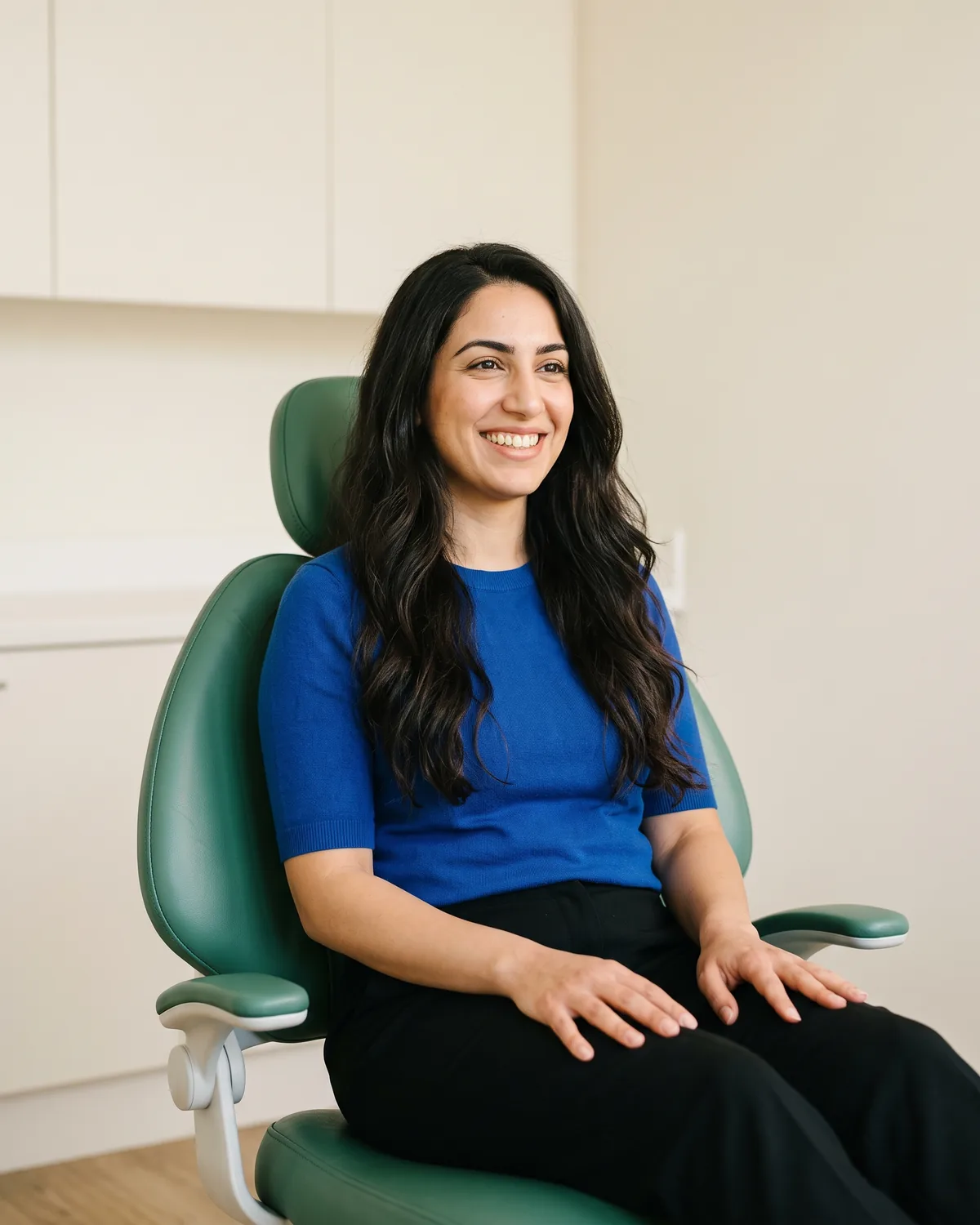 Woman with long dark waves and a blue top seated in a green dental chair.