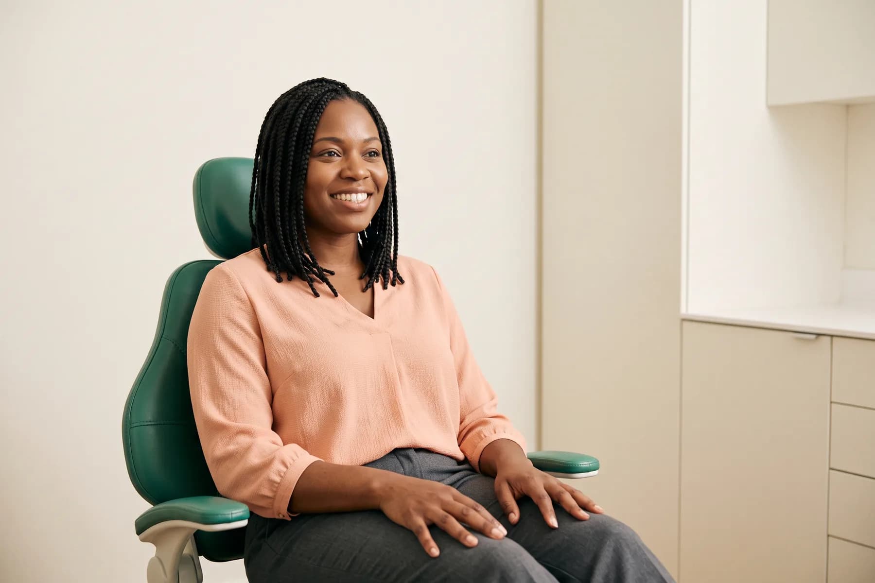Woman with shoulder-length braids and a peach blouse smiling in a green dental chair.