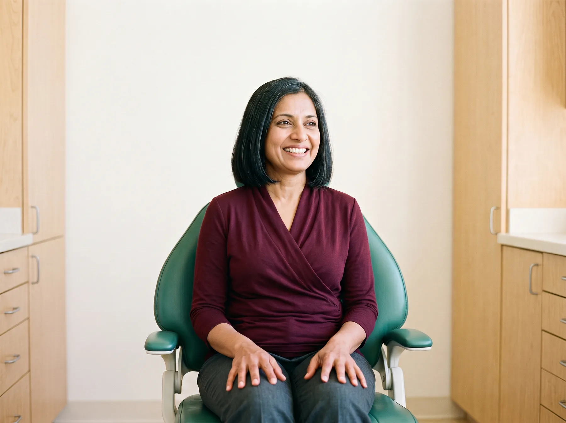 Woman with a black bob and a burgundy top smiling in a green dental chair.