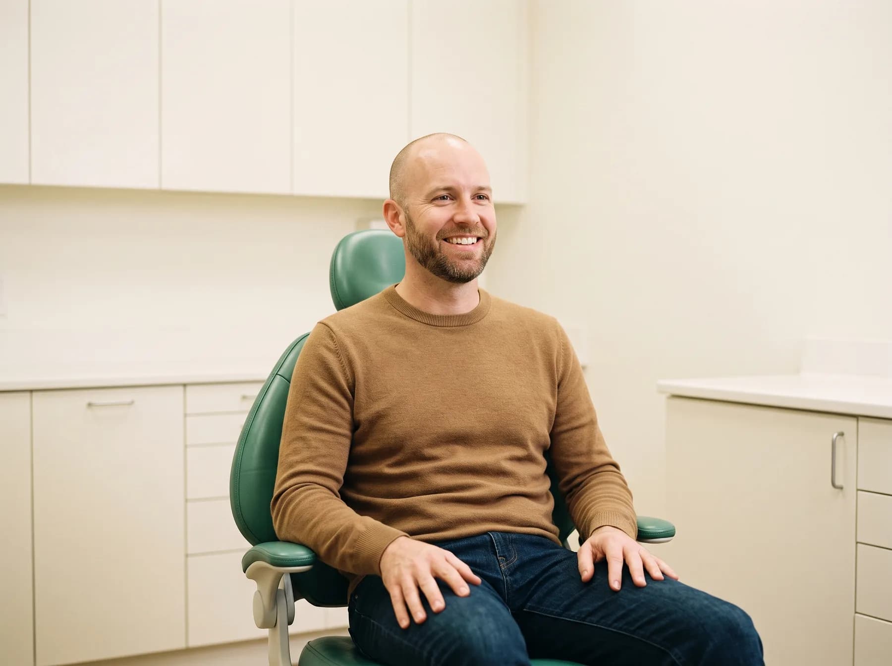 Bald man with a short beard and a camel sweater smiling in a green dental chair.