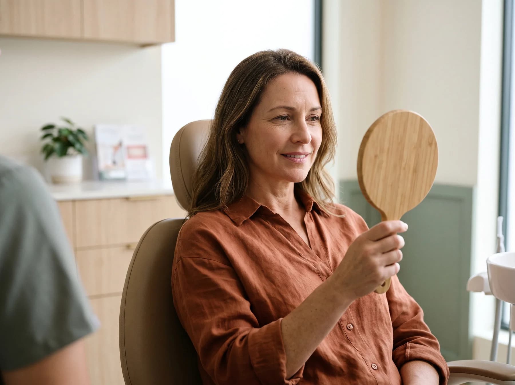 Illustrative portrait of a woman looking into a handheld mirror in a consultation room.