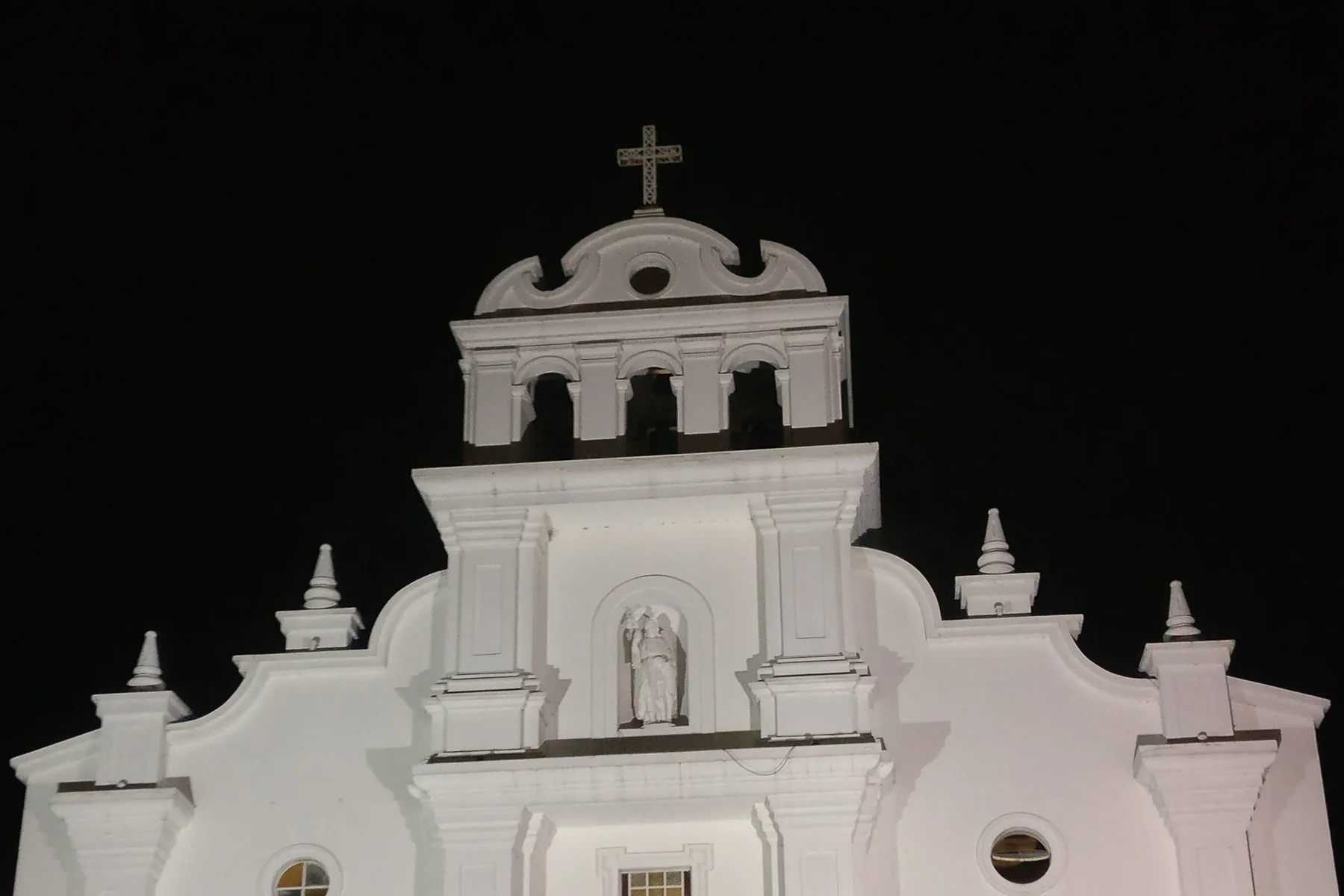 An upper facade detail of Iglesia San Rafael shows its cross, bells and sculpture at night in Escazú, Costa Rica.